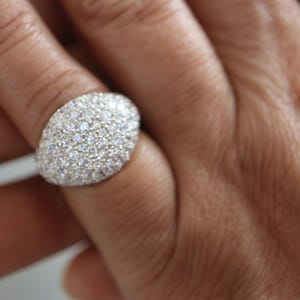May include: A dome-shaped ring featuring a dense pavé setting of numerous small, clear, round-cut stones that cover the entire visible surface of the band. The metal setting appears silver in color, providing a bright, reflective base for the stones.