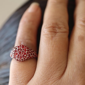 May include: A silver ring with a rounded top, densely covered in small, round, red gemstones. The ring is worn on a finger, showcasing the vibrant red color and intricate design. The ring's surface is smooth and polished.