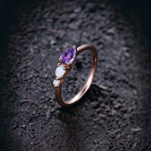 May include: A delicate rose gold ring with a pear-shaped amethyst gemstone, a round opal gemstone, and a small diamond accent. The ring is set on a dark textured background.