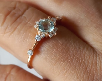 May include: A delicate gold ring with a light blue gemstone surrounded by smaller clear gemstones. The ring is on a finger.