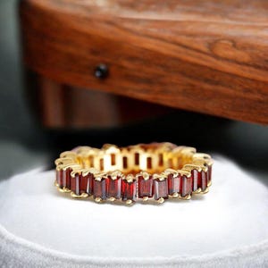 May include: A gold band ring with a row of red rectangular gemstones. The ring is on a white surface.