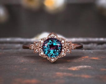 May include: A rose gold engagement ring with a round, blue-green alexandrite gemstone surrounded by a halo of smaller diamonds. The ring features a delicate floral design with leaf accents.