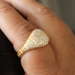 May include: A gold ring with a teardrop-shaped face, encrusted with small, sparkling white gemstones. The ring is on a finger, showcasing the intricate details of the jewelry. The band has a textured design.