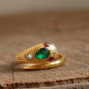 May include: A gold-colored ring in the shape of a snake, with a textured surface. The snake's head features a large green gemstone, surrounded by smaller red and clear gemstones. The ring is set against a wooden background.
