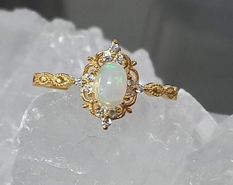 May include: A gold ring with a white opal gemstone set in a filigree design. The ring is surrounded by small clear stones.
