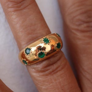 Puede incluir: Un anillo de oro con una banda ancha y una fila de piedras preciosas de color verde esmeralda engastadas en la banda.