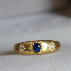 May include: A gold ring with three gemstones. The center stone is a dark blue oval, flanked by two smaller, clear stones. The ring has a starburst design around each stone.