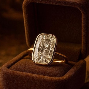 May include: A gold ring with a large, rectangular, clear gemstone. The gemstone is set in a gold bezel and has intricate facets. The ring is displayed in a brown velvet ring box, creating a luxurious presentation.