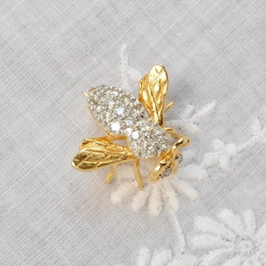 May include: A gold-toned bee brooch with a body encrusted with sparkling clear crystals. The wings and legs are detailed with textured gold. The brooch is set against a white, textured background.