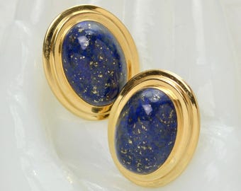 Pode incluir: Um par de brincos ovais com cabochões de lápis-lazúli polidos com manchas naturais de cor dourada. Cada pedra está engastada numa moldura de metal de tom dourado lisa e arredondada, criando um design clássico e simétrico para estes acessórios.