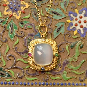 May include: A gold pendant necklace featuring a large, smooth, translucent gray cabochon stone set in an ornate gold frame. The frame is accented with a small clear stone at the top and three small round stones in pink, green, and blue.