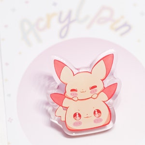 Könnte beinhalten: Eine rosa und weiße Acryl-Anstecknadel mit zwei Comicfiguren. Die obere Figur ist ein rosa Fuchs mit weißem Bauch und die untere Figur ist ein weißer Fuchs mit rosa Bauch. Beide Figuren haben geschlossene Augen und lächeln.