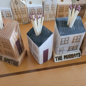 May include: Five wooden matchstick holders shaped like houses with black roofs. Each house has a different name on the base: The Smiths, The Murrays, and three others. The houses are arranged in a row on a wooden surface.