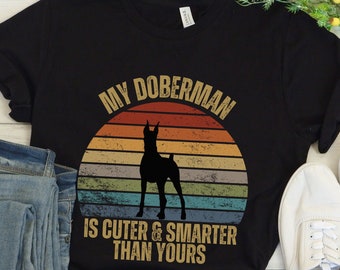 Op de afbeelding: Zwart T-shirt met een vintage-stijl afbeelding van een Dobermann. De afbeelding zegt "Mijn Dobermann is schattiger & slimmer dan die van jou".