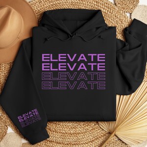 Könnte beinhalten: Schwarzer Hoodie mit einem viermal wiederholten lilafarbenen "ELEVATE"-Motiv. Der Hoodie hat eine Kapuze mit Kordelzug und lange Ärmel.