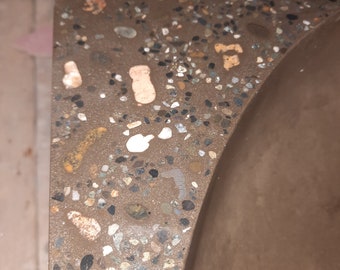 Pode incluir: Close-up de uma bancada de concreto marrom com pequenas manchas coloridas de agregado. A bancada tem uma borda curva e um acabamento liso e polido.