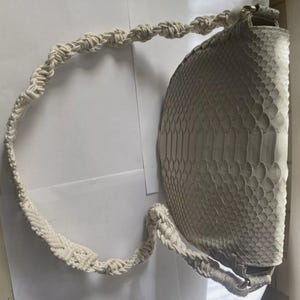 May include: A white snakeskin handbag with a white crocheted strap. The bag has a rounded shape and a textured surface.