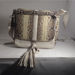May include: A cream-colored crossbody bag with a flap closure, featuring a snakeskin pattern in shades of brown and beige. The bag has a cream-colored leather strap and a tassel detail. The bag is approximately 10 inches wide.