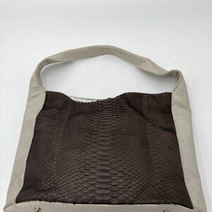 May include: A light beige leather handbag with a brown snakeskin panel. The bag has a single strap and two silver metal accents.