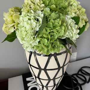May include: A decorative vase filled with green and light green hydrangeas. The vase is cream-colored with a black geometric pattern. The vase sits on a white shelf with a black book that says "TOM" on the cover.