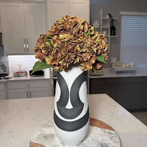 May include: A white vase with a black geometric design holds a bouquet of brown and yellow flowers with green leaves. The vase sits on a marble and wood decorative tray, suitable for home decor.