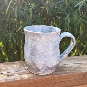 May include: A handmade ceramic mug with a light blue and gray glaze. The mug has a rounded body, a sturdy handle, and a textured surface. The mug is sitting on a wooden surface with a green background.