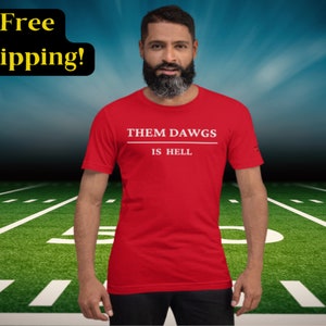 May include: Red t-shirt with white text that reads "Them Dawgs Is Hell". A man is wearing the shirt and standing in front of a green football field.