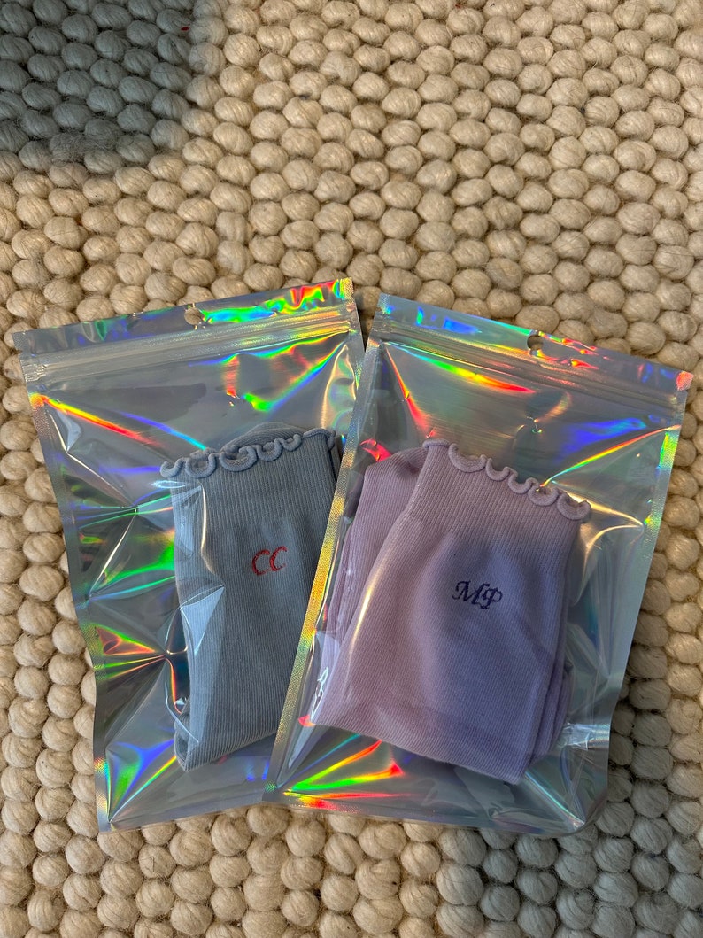 May include: Two pairs of socks in individual clear plastic bags with iridescent rainbow patterns. The socks are both ribbed and have a frilly edge. One pair is light blue with the initials "CC" embroidered in red. The other pair is lavender with the initials "MP" embroidered in black.