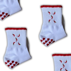 May include: White socks with red trim, featuring red chilli pepper and heart designs. The socks have a ribbed texture and are decorated with small red chilli peppers and red heart-shaped grips on the soles.