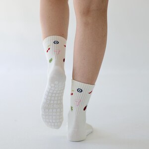 May include: Cream-coloured socks featuring embroidered patterns of a blue eye, red chilli, and a pink mushroom. The soles have a non-slip grip. These socks are a comfortable, everyday accessory.