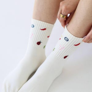 May include: A pair of white socks with colourful embroidered designs. The socks feature small illustrations of a red chilli pepper, a ladybug, a blue eye, and other small shapes. The socks are pulled up on the legs.