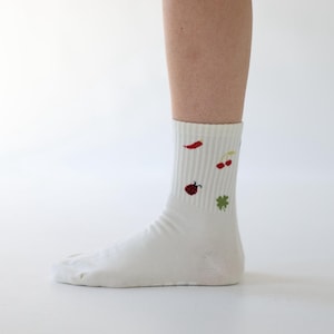 May include: White socks featuring embroidered motifs: a red chilli, cherries, a ladybird, and a four-leaf clover. The socks have a ribbed cuff and are worn on a leg against a white backdrop.
