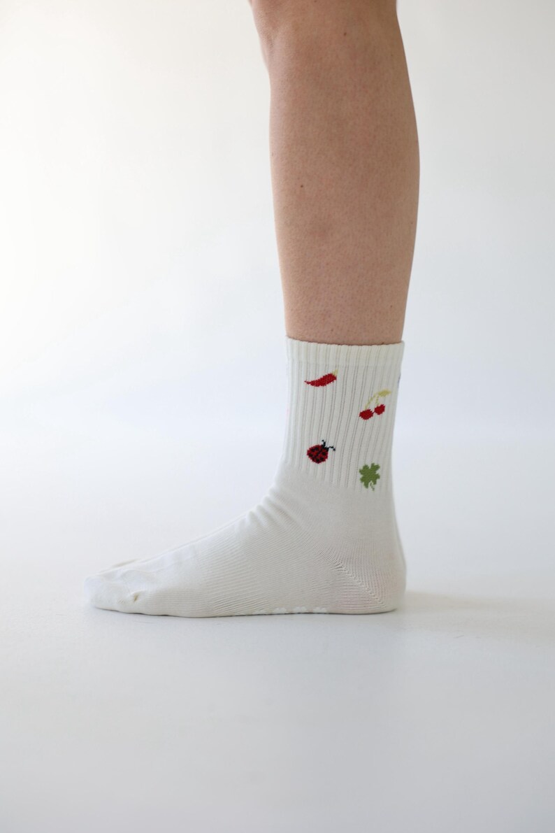 May include: White socks featuring embroidered motifs: a red chilli, cherries, a ladybird, and a four-leaf clover. The socks have a ribbed cuff and are worn on a leg against a white backdrop.