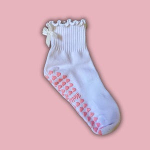 May include: White ankle socks with a pink heart pattern on the bottom and a white bow on the top. The socks have a frilly edge.