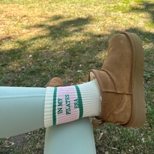 May include: White socks with green and pink stripes and the text "In My Pilates Era" on them. The socks are worn on a person's feet with brown suede boots.