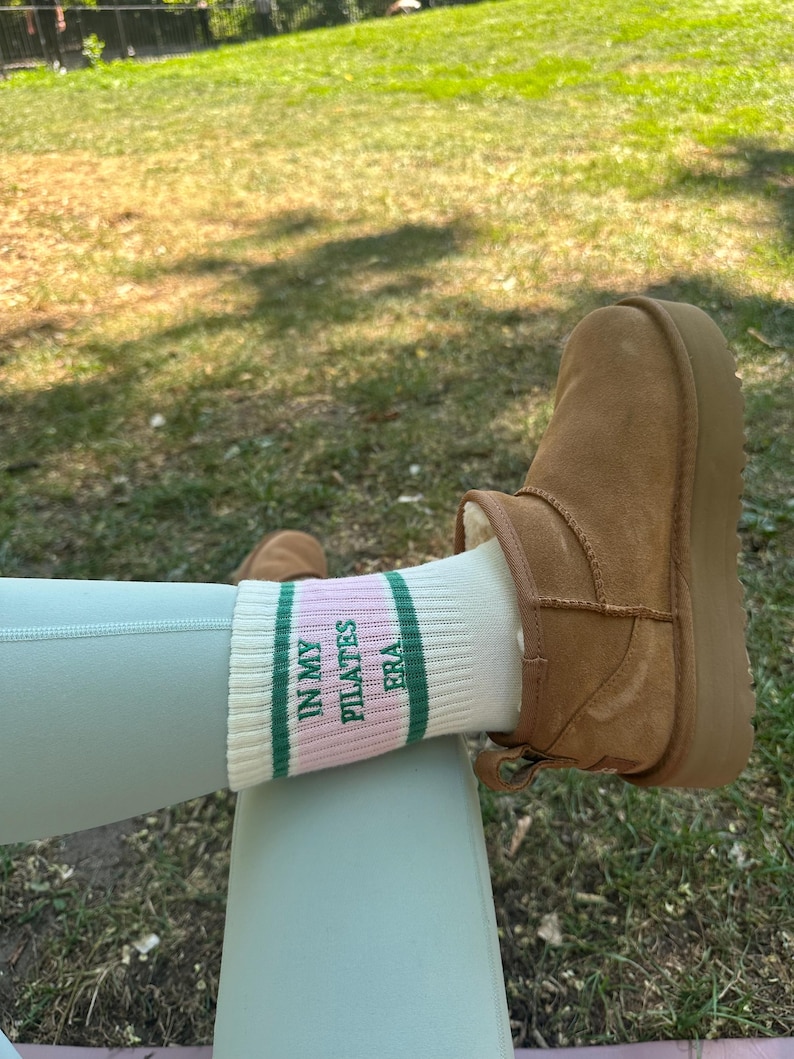 May include: White socks with green and pink stripes and the text "In My Pilates Era" on them. The socks are worn on a person's feet with brown suede boots.