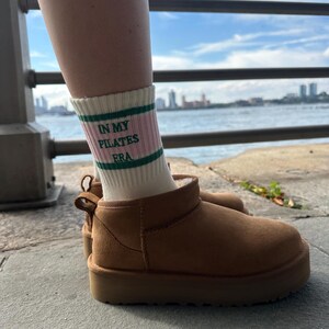 May include: A pair of brown suede ankle boots with a white sock that reads "In My Pilates Era" in green letters. The boots are worn by a person standing on a concrete surface.