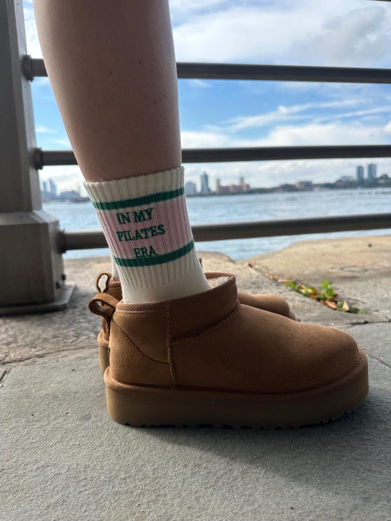 May include: A pair of brown suede ankle boots with a white sock that reads "In My Pilates Era" in green letters. The boots are worn by a person standing on a concrete surface.