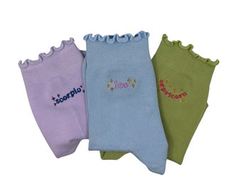 May include: Three pairs of colorful socks with embroidered zodiac signs. The socks are light purple, light blue, and light green. The signs are "Scorpio", "Leo", and "Capricorn".