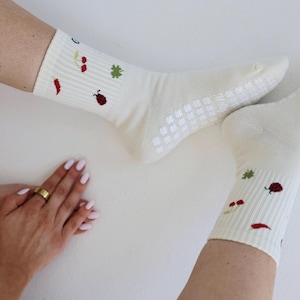 May include: Cream-coloured socks with a ribbed pattern and small embroidered motifs of a ladybug, chilli pepper, and other plants. The socks have a non-slip grip on the soles. A hand with a gold ring is visible.