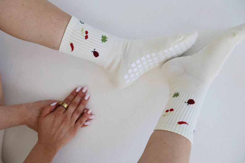 May include: Cream-coloured socks with a ribbed pattern and small embroidered motifs of a ladybug, chilli pepper, and other plants. The socks have a non-slip grip on the soles. A hand with a gold ring is visible.