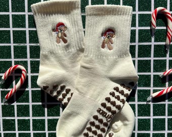 Puede incluir: Un par de calcetines color crema con un patrón de corazones marrones en las suelas y un pequeño oso de peluche bordado con un gorro de Papá Noel. La palabra "Grace" está impresa en la suela. Bastones de caramelo esparcidos.