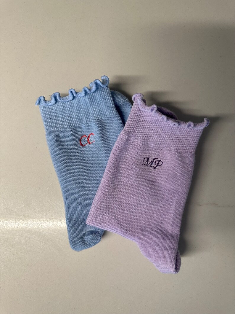 May include: Two pairs of socks with frilly tops. One pair is light blue with the initials "CC" embroidered on them. The other pair is lavender with the initials "MP" embroidered on them.