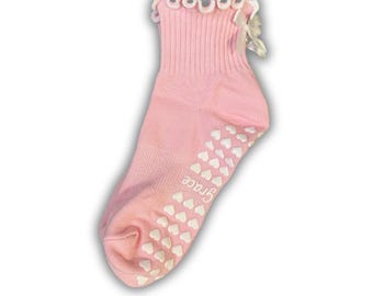 May include: Pink baby socks with white heart-shaped grips and a white bow on the ankle. The socks have a white lace trim and the word "Grace" is printed on the bottom.