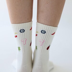 May include: A pair of cream-coloured socks with colourful embroidered designs. The socks feature a pattern of eyes, cherries, chilli peppers, clovers, and ladybirds. The socks are ribbed and reach mid-calf.