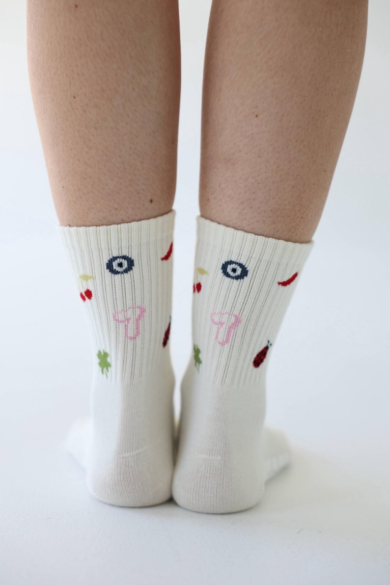 May include: A pair of cream-coloured socks with colourful embroidered designs. The socks feature a pattern of eyes, cherries, chilli peppers, clovers, and ladybirds. The socks are ribbed and reach mid-calf.