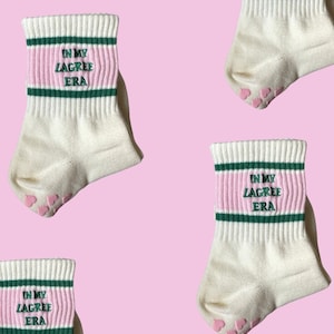 May include: Four pairs of white socks with pink and green stripes. The socks have the text "In My Lagree Era" embroidered on them. The socks have pink heart-shaped grips on the bottom.