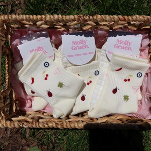 May include: A woven basket containing several pairs of cream-coloured socks. Each sock has embroidered designs of cherries, ladybirds, and clovers. The socks are packaged with tags that read "Molly Grace."