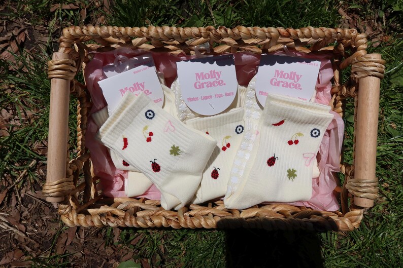 May include: A woven basket containing several pairs of cream-coloured socks. Each sock has embroidered designs of cherries, ladybirds, and clovers. The socks are packaged with tags that read "Molly Grace."