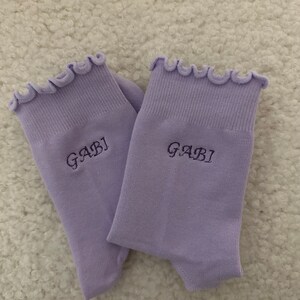 May include: A pair of lavender socks with a frilly top edge. The socks have the name "GABI" embroidered on them.